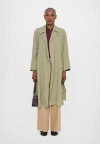 Cappotto lungo verde oliva con maniche larghe, abbinato a una polo bordeaux e a pantaloni beige a gamba larga. Una borsa nera e scarpe completano il look.