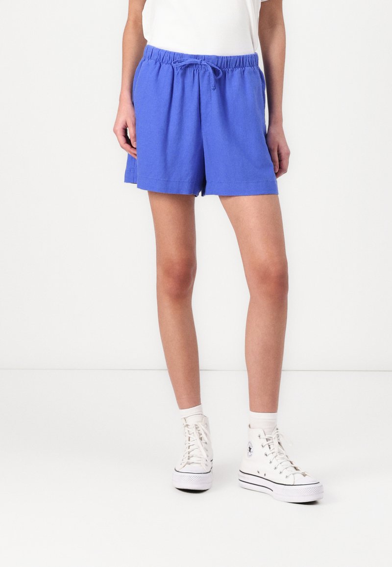Pantalones cortos azules de cortes relajados con cintura elástica y cordón. Combinados con zapatillas blancas de caña alta y calcetines tobilleros. Diseño simple, sin patrones.