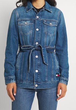 Femme portant une veste en jean bleue avec boutons sur le devant, ceinture à la taille, poches poitrine, et un patch du logo Tommy Jeans sur le côté inférieur.