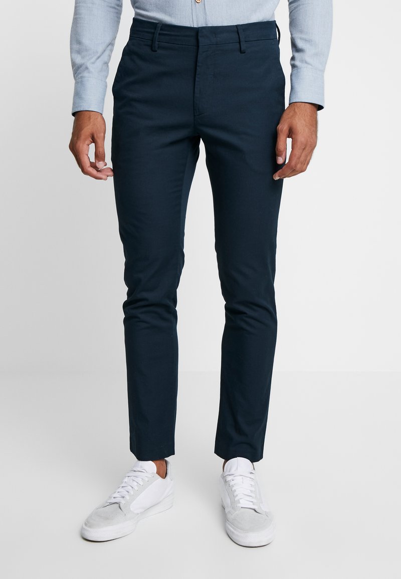 Calças de algodão azul-marinho com um corte Slim, apresentando passantes para cinto e bolsos laterais, combinadas com ténis cinza-claro.