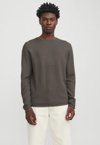 Camiseta de manga larga texturizada en marrón con cuello redondo, que presenta un patrón tejido y mangas ajustadas, combinada con pantalones de color claro.