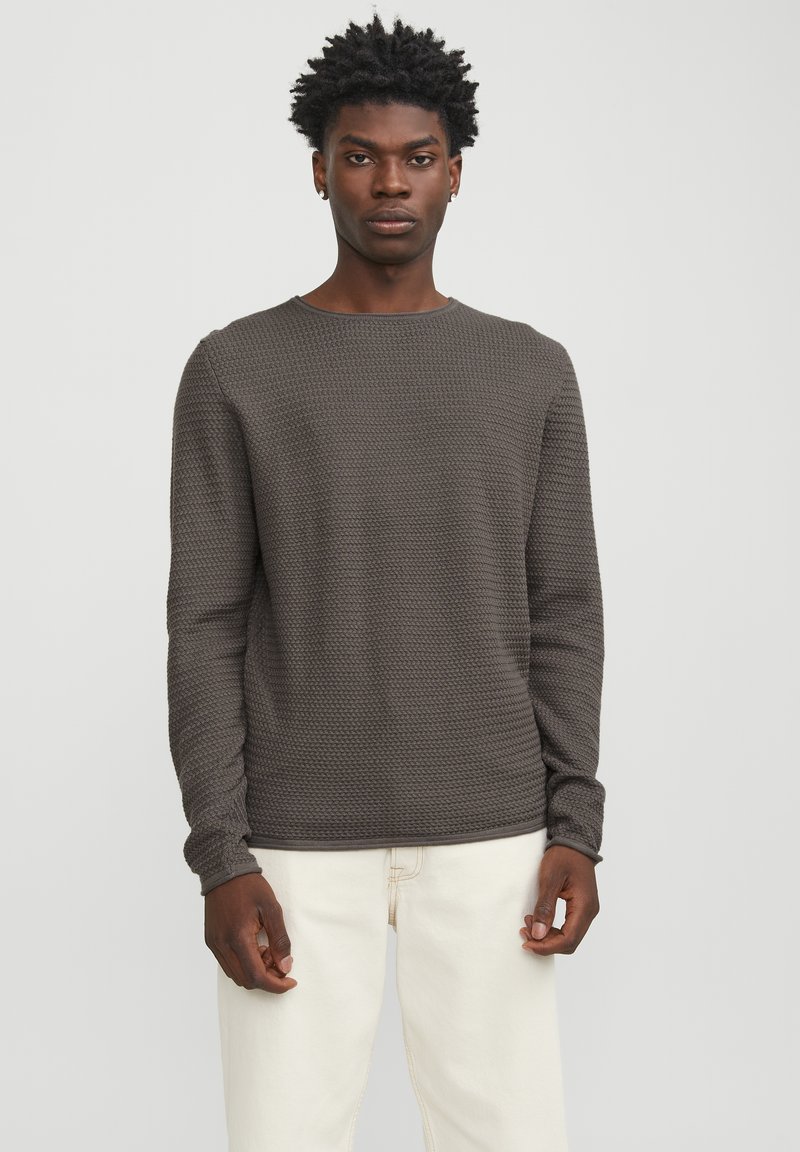 Camiseta de manga larga texturizada en marrón con cuello redondo, que presenta un patrón tejido y mangas ajustadas, combinada con pantalones de color claro.