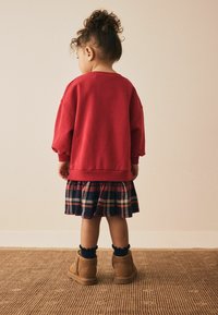 Roter Sweatshirt mit lockeren Ärmeln, über einem karierten Rock in Marineblau, Rot und Gelb geschichtet. Braune Stiefel mit weicher Textur und gerüschten marineblauen Socken.