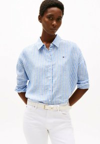 Hellblau-weiß gestreifte Button-Up-Hemd mit Kragen, hochgekrempelten Ärmeln und kleinem Logo, kombiniert mit weißen Hosen und einem weißen Gürtel.