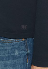 Námornícky pletený sveter so subtílnym logom "50" na leme, v kombinácii s modrými džínsami s viditeľným gombíkom na vrecku.