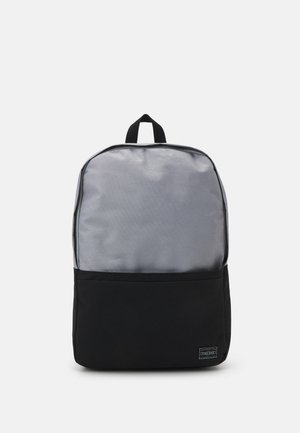 Šedý látkový batoh s hladkou horní částí a texturovanou černou spodní částí. Obsahuje vrchní držadlo a výraznou značku na přední straně.