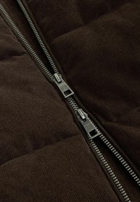 Brun fløjlspuffer vest med en sølvlynlås; har et quiltet mønster og dobbelte lynlåsdrag, der fremhæver dets funktionelle design.