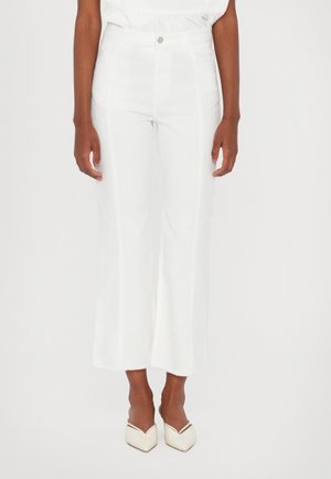 Vrouw draagt witte hooggetailleerde cropped broek met een knoop en twee voorzakken, gecombineerd met witte pumps met puntige neus.