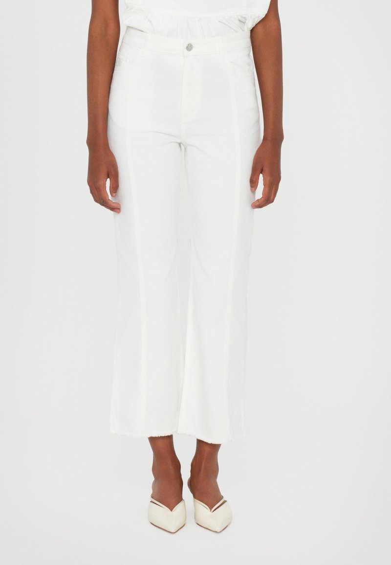 Femme portant un pantalon blanc court taille haute avec un bouton et deux poches avant, associé à des talons blancs à bout pointu.