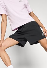 Fekete atlétikai rövidnadrág elasztikus derékpánttal, rajta szürke Adidas logó. Világos rózsaszín pólóval párosítva. Sima anyagtextúra.