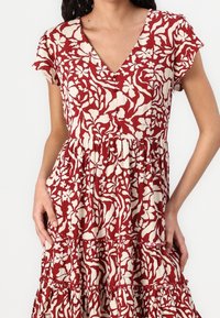 Femme portant une robe à manches courtes avec un motif floral rouge et blanc, boutonnée sur le devant, tenant la jupe avec les deux mains.