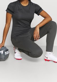 Sort kortærmet atletisk t-shirt kombineret med mørkegrå bukser. En grå fodbold ligger i nærheden. Fodtøjet er hvidt med røde detaljer og en Nike-logo.