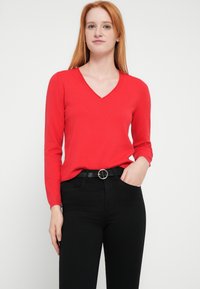 Suéter rojo de manga larga con cuello en V, fabricado con tela suave, combinado con jeans negros y un cinturón negro con una hebilla redonda plateada.