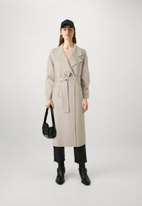 Cappotto di lana grigio chiaro con vita cinturata, ampie rever, e dettagli con bottoni, abbinato a stivali neri alla caviglia e una piccola borsa nera.