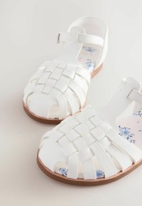 Weiße geflochtene Sandalen mit offener Zehe und Knöchelriemen. Die leicht gepolsterte Innensohle zeigt ein florales Muster in Blau. Strapazierfähige braune Sohle.