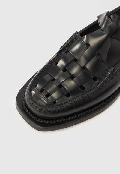Μαύρο δερμάτινο παπούτσι με υφαντό σχέδιο στο επάνω μέρος, επίπεδη σόλα και διακοσμητική ραφή κατά μήκος των άκρων. Στρογγυλερό σχήμα μύτης.