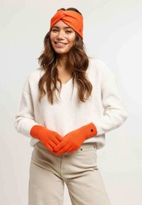 Gants tricotés orange et bandeau, avec un design texturé et un détail torsadé, associés à un pull à côtes blanc et un pantalon beige.