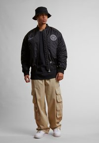 Jachetă bomber neagră cu un logo circular, purtată peste un tricou negru. Pantaloni cargo bej cu multiple buzunare, asortați cu adidași albi. Pălărie bucket neagră.