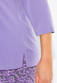 Lavendelfarbenes Kurzarmoberteil mit seitlichem Schlitz, aus weichem Stoff gefertigt. Verfügt über eine dezente Textur und eine Nahtdetails entlang der Ärmel und des Saums.