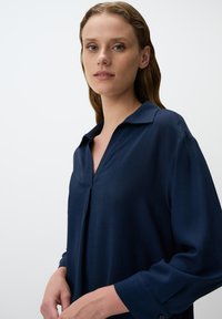 Blusa blu navy con colletto e una sottile piega nella parte anteriore. Realizzata in tessuto morbido, con maniche lunghe e polsini.