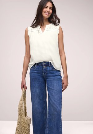 Femme souriante, portant un haut blanc sans manches avec détail œillet, jean bleu à jambes larges, tenant un sac fourre-tout tissé beige sur fond uni.