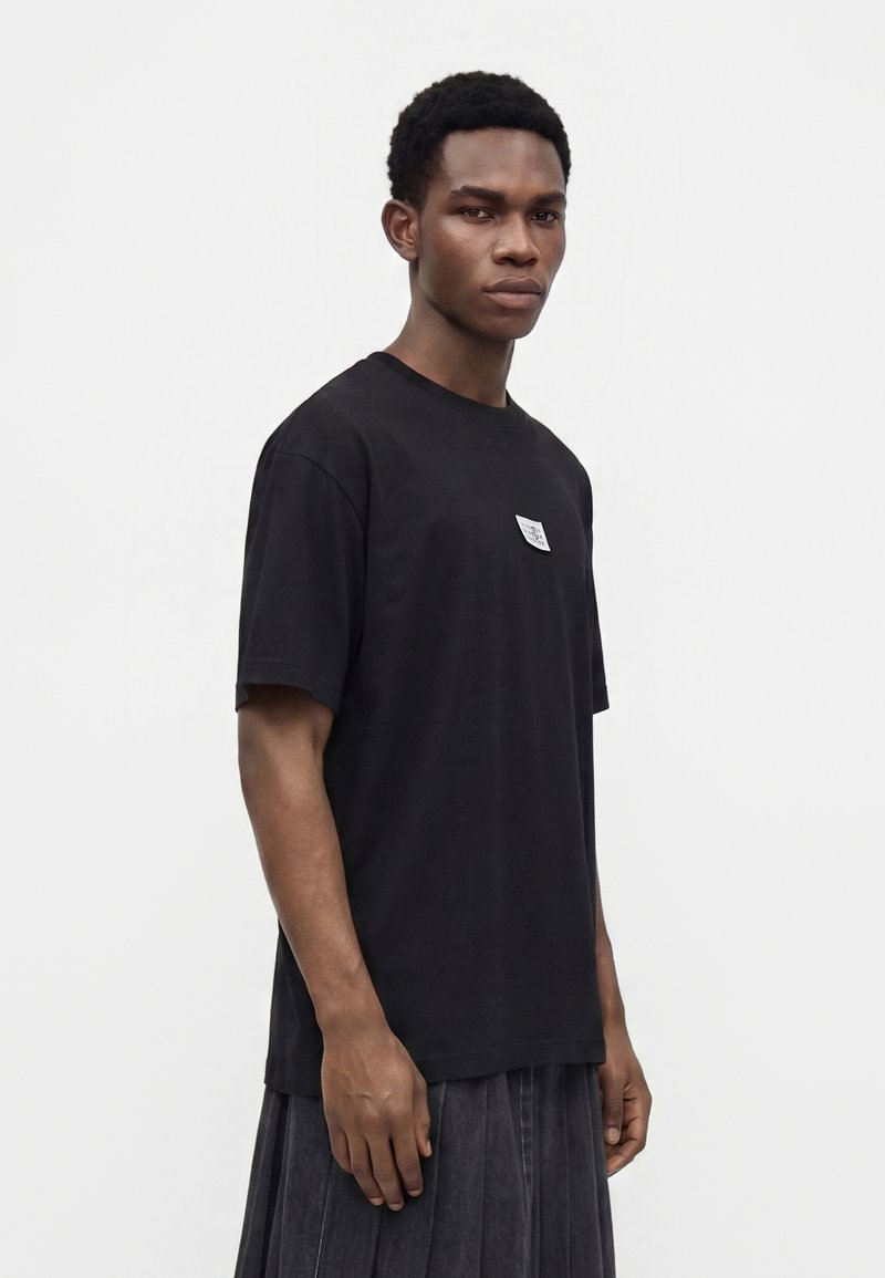 Joven con cabello corto y rizado, vestido con una camiseta negra y pantalones plisados oscuros sobre un fondo claro.