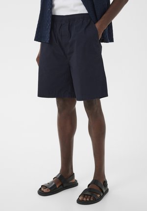 Homme portant un short bleu marine à taille élastique avec poches latérales et des sandales noires à bride ouverte, debout avec une main dans la poche.