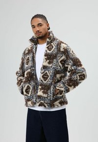 Zachte jas met bruin- en crèmekleurig patroon en blauwe accenten, hoge kraag, voorzakken en een ritssluiting, gedragen over een wit T-shirt.