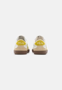 Fehér sneaker sárga akcentusokkal és bézs velúr panelekben. Gumi talppal rendelkezik, amely textúrázott mintázattal készült, és "VEJA" márkajelzéssel a sarok részen.