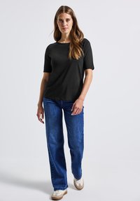 Femme se tenant debout portant un haut noir à manches courtes, un jean bleu et des baskets blanches devant un fond clair.