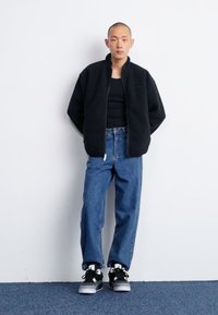 Giacca con zip in pile nera sopra una canotta nera, abbinata a jeans larghi blu chiaro e sneakers nere con dettagli bianchi.