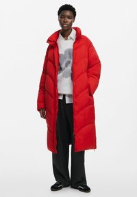 Abrigo acolchado rojo de diseño largo, con secciones acolchadas, un cuello alto y botones a presión. Lleva puesto un suéter gráfico gris y pantalones negros.