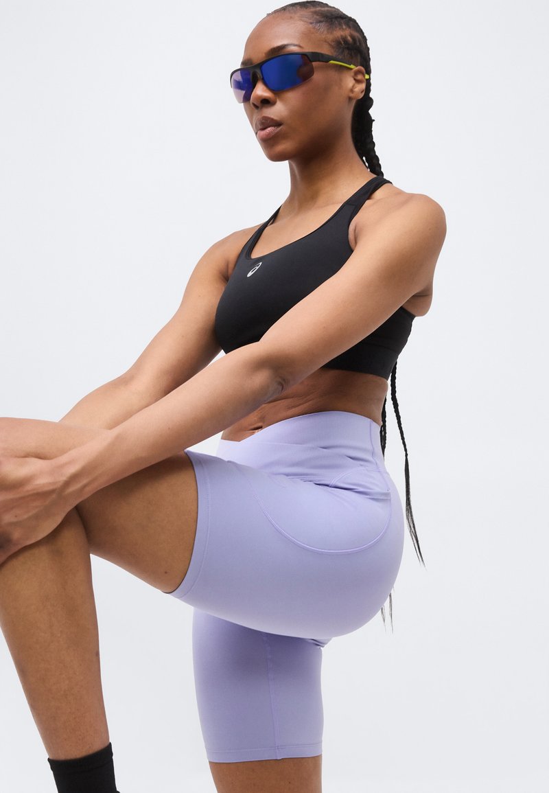 Vrouw in een zwart sportbeha, lichtpaarse fietsbroek en blauwgetinte zonnebril strekt haar been en houdt haar knie met beide handen vast.