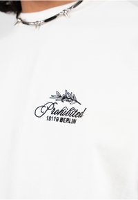 Hvit t-skjorte med svart brodert tekst: "Forbudt 10119 BERLIN" med et bladdesign. Mykt bomullsmateriale. Klassisk rund halsform.