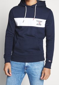 Mężczyzna w granatowym hoodie z białym paskiem na klatce piersiowej i logo "Tommy Jeans", w połączeniu z jasnoniebieskimi dżinsami.