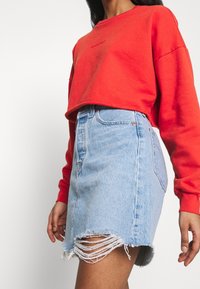 Sudadera corta roja con cuello redondo, combinada con una mini falda de mezclilla azul clara que presenta un dobladillo deshilachado y detalles desgastados.