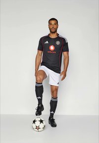 Juodas futbolo marškinėlis su baltais akcentais ir logotipais, derinamas su baltais šortais ir juodomis kojinėmis. Žaidėjas stovi ant futbolo kamuolio.