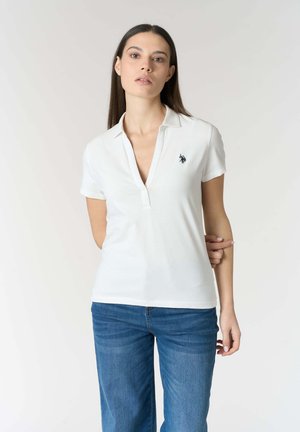 Donna con capelli lunghi e scuri che indossa una polo bianca e jeans blu, in piedi davanti a uno sfondo chiaro e semplice.