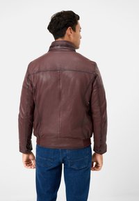 Brun läderjacka med hög krage, ribbade ärmslut och sömmar vid ryggen. Buren med blå denimjeans. Slät textur, strukturerad design.
