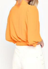 Camicetta arancione corta con maniche a sbuffo, orlo elastico e texture liscia, abbinata a pantaloni bianchi a vita alta.