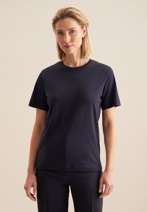 Femme aux cheveux blonds courts portant un t-shirt bleu marine uni à manches courtes et un pantalon foncé assorti sur un fond beige.
