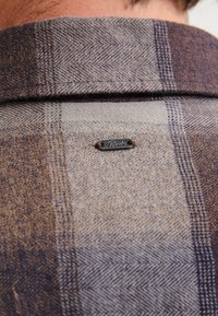 Colletto di camicia a quadri in toni di marrone e grigio, con un piccolo tag metallico con logo. Il tessuto appare testurizzato con motivi contrastanti.