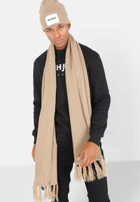 Beige gebreide sjaal met gefranjerde uiteinden over een zwarte trui, gedragen door een persoon in een beige beanie met een wit logo label.