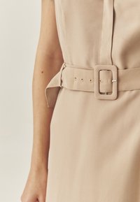Beiges Kleid mit tailliertem Design, das einen breiten Gürtel mit quadrischer Schnalle aufweist, aus glattem Stoff gefertigt, mit einem Knopfdetail an der Taille.