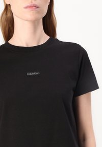 Tricou din bumbac negru, cu mâneci scurte, guler rotund și o etichetă mică neagră cu "Calvin Klein" pe piept. Textură netedă.