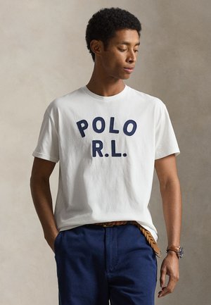 Fehér "POLO R.L." pólót, tengerészkék nadrágot, barna fonott övet és karórát viselő férfi, zsebeiben tartja a kezét, egyszínű háttér előtt áll.