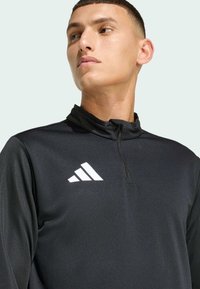 Μαύρος αθλητικός πόλο με ψηλό γιακά, μπροστινό μισό φερμουάρ και λευκό λογότυπο Adidas με τρεις ρίγες στο αριστερό στήθος. Απαλή, ελαφριά ύφανση.