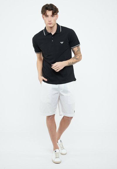 Jonge man met krullend haar, gekleed in een zwart poloshirt, witte shorts, witte sneakers en zichtbare armtattoos, staande tegen een witte achtergrond.