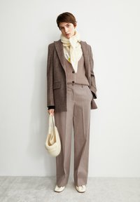Bruine oversized blazer met een gestructureerde afwerking, beige gebreid trui, brede taupe broek, witte gebreide sjaal en een crèmekleurige tote bag.