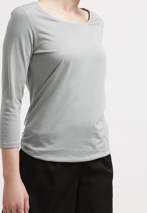 Person trägt ein hellgraues, langärmeliges Shirt mit rundem Ausschnitt und schwarze Hose vor einem einfarbigen weißen Hintergrund.